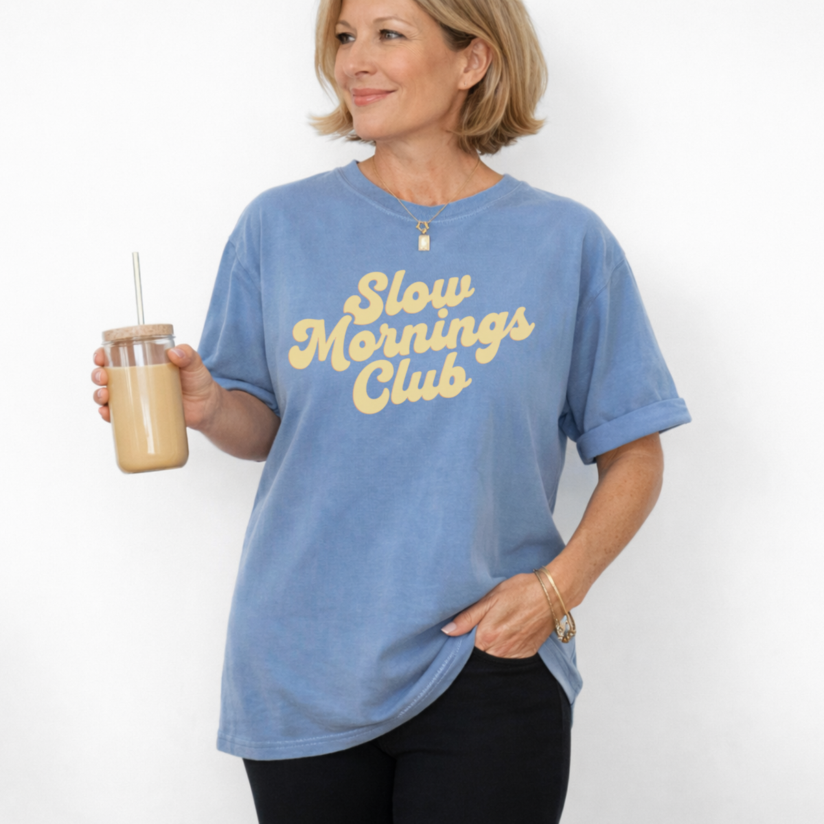 Woman wearing a blue t-shirt with 'Slow Mornings Club' text, holding a drink.