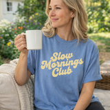 Woman wearing a blue 'Slow Mornings Club' t-shirt holding a mug outdoors.