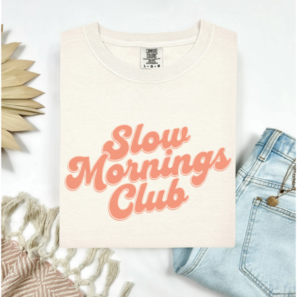 Ivory t-shirt with 'Slow Mornings Club' text on a white background with denim jeans and decorative elements.