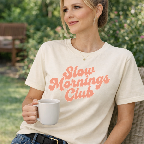 Woman wearing a 'Slow Mornings Club' t-shirt holding a mug outdoors.