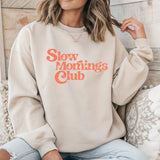 Woman wearing a beige crewneck sweatshirt with 'Slow mornings club' text in orange.