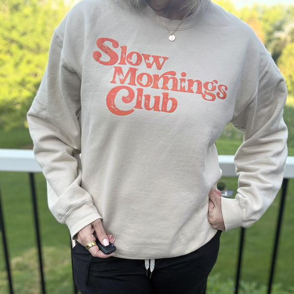 A person wearing a cream-colored crewneck sweatshirt with 'Slow Mornings Club' printed in a distressed coral-red font across the chest, paired with black pants, standing outdoors in front of a railing and green foliage.