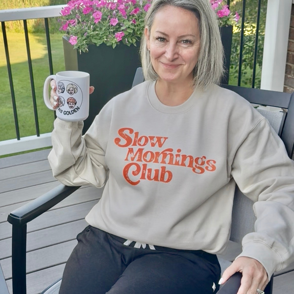 A woman sitting on a patio chair holding a white mug, wearing a beige sweatshirt with 'Slow Mornings Club' printed in orange text. The background shows a wooden deck, green grass, and pink flowers.