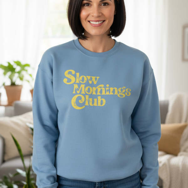 Woman wearing a blue sweatshirt with 'Slow Mornings Club' text in a living room setting