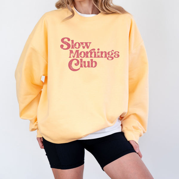 Woman wearing a yellow sweatshirt with 'Slow Mornings Club' pink text on a plain background
