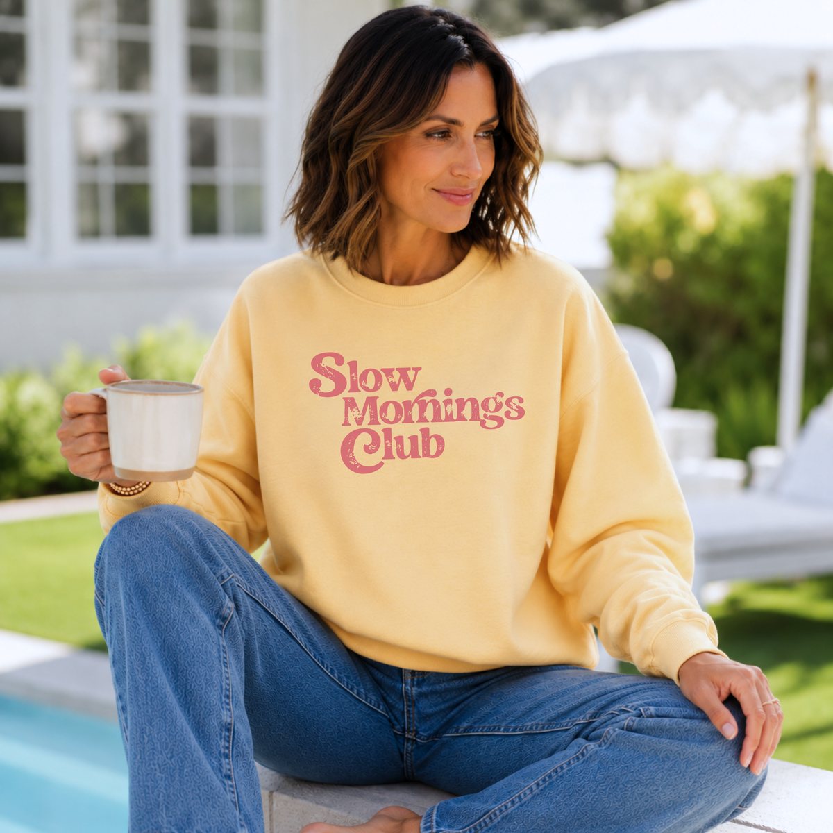 Woman wearing a yellow sweatshirt with 'Slow Mornings Club' text, sitting by a pool.