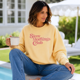 Woman wearing a yellow sweatshirt with 'Slow Mornings Club' text, sitting by a pool.