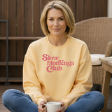 Woman wearing a yellow sweatshirt with 'Slow Mornings Club' text, holding a mug outdoors.