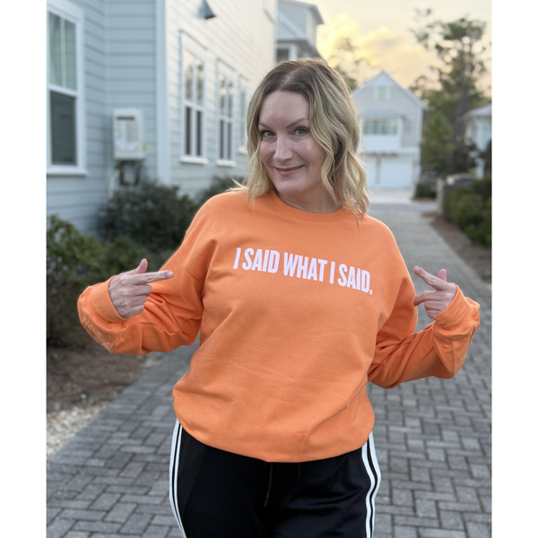 Woman standing outside smiling with an tangerine orange sweatshirt on pointing to the words on her sweatshirt that say "I said what I said."