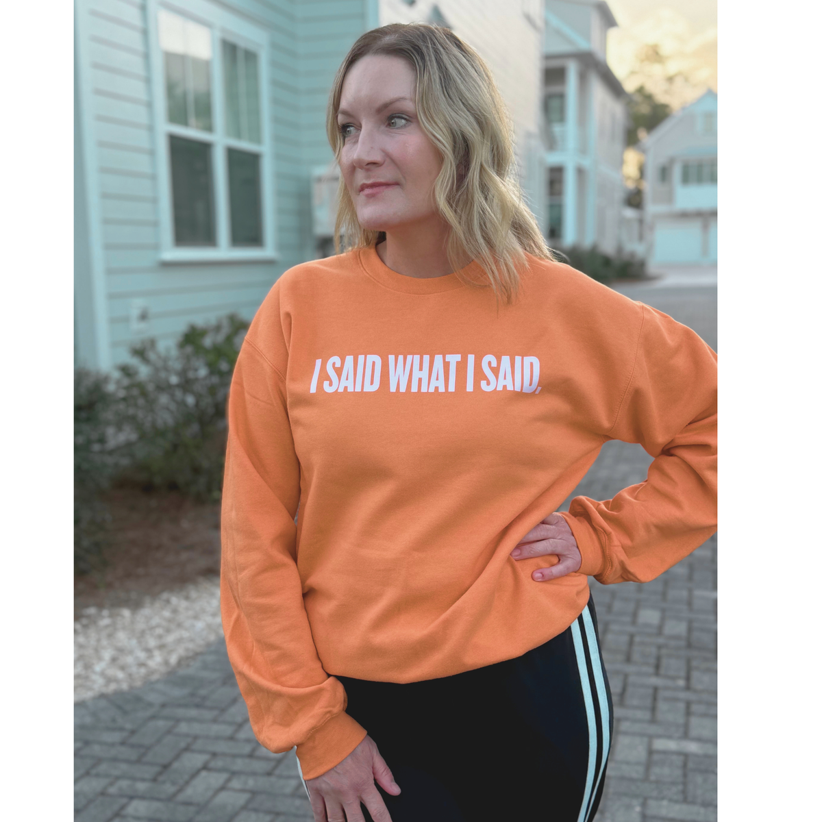 Woman wearing an orange sweatshirt with text reading "I said what I said," in pink font standing outdoors.