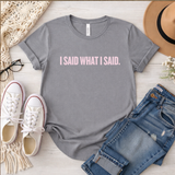 Gray t-shirt with 'I SAID WHAT I SAID' text, paired with jeans and sneakers on a light wooden surface.