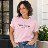 A woman wearing a light pink crewneck t-shirt with the text 'sarcasm IS MY LOVE LANGUAGE' printed in dark green on the front, paired with blue jeans, standing in front of a white house exterior.