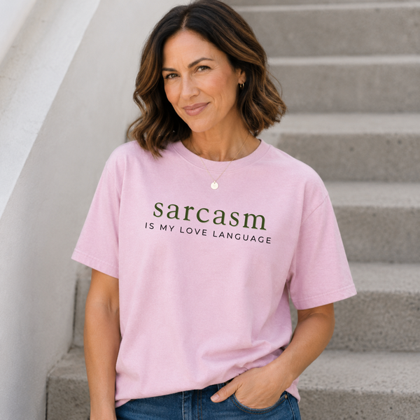 A woman wearing a light pink graphic t-shirt with the text 'sarcasm IS MY LOVE LANGUAGE' printed in dark green and black, standing on concrete stairs.