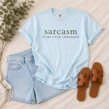A light blue graphic T-shirt laid flat on a beige fabric surface, styled with light blue denim shorts, brown slide sandals, and eucalyptus branches. The shirt has dark green and black text reading 'sarcasm IS MY LOVE LANGUAGE'.