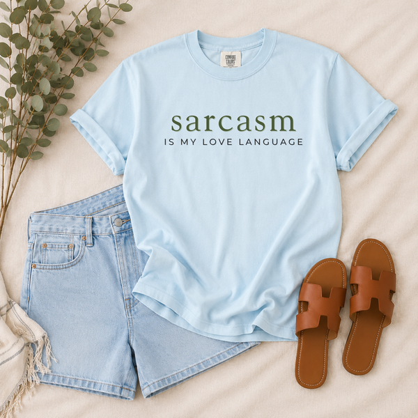 A light blue graphic T-shirt laid flat on a beige fabric surface, styled with light blue denim shorts, brown slide sandals, and eucalyptus branches. The shirt has dark green and black text reading 'sarcasm IS MY LOVE LANGUAGE'.