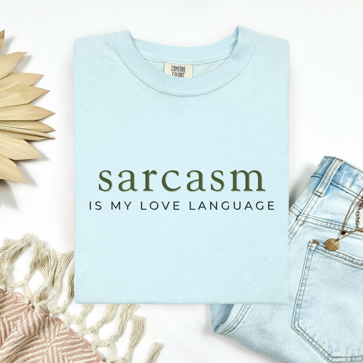 A folded light blue t-shirt with the text 'sarcasm IS MY LOVE LANGUAGE' printed on the front, styled in a flat lay with light-wash jeans, a woven textile, and a decorative wooden sun on a white background.