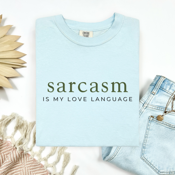 A folded light blue t-shirt with the text 'sarcasm IS MY LOVE LANGUAGE' printed on the front, styled in a flat lay with light-wash jeans, a woven textile, and a decorative wooden sun on a white background.