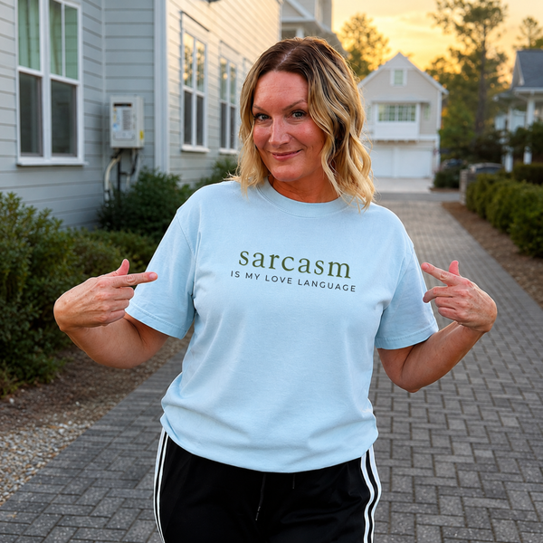 A woman stands on a paved walkway in front of suburban houses, wearing a light blue graphic T-shirt that reads 'sarcasm IS MY LOVE LANGUAGE' in dark green text, paired with black track pants with white side stripes.