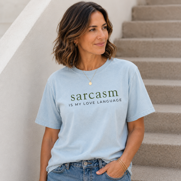 A woman wearing a light blue graphic t-shirt with the text 'sarcasm IS MY LOVE LANGUAGE' printed in dark green, standing on outdoor concrete stairs.