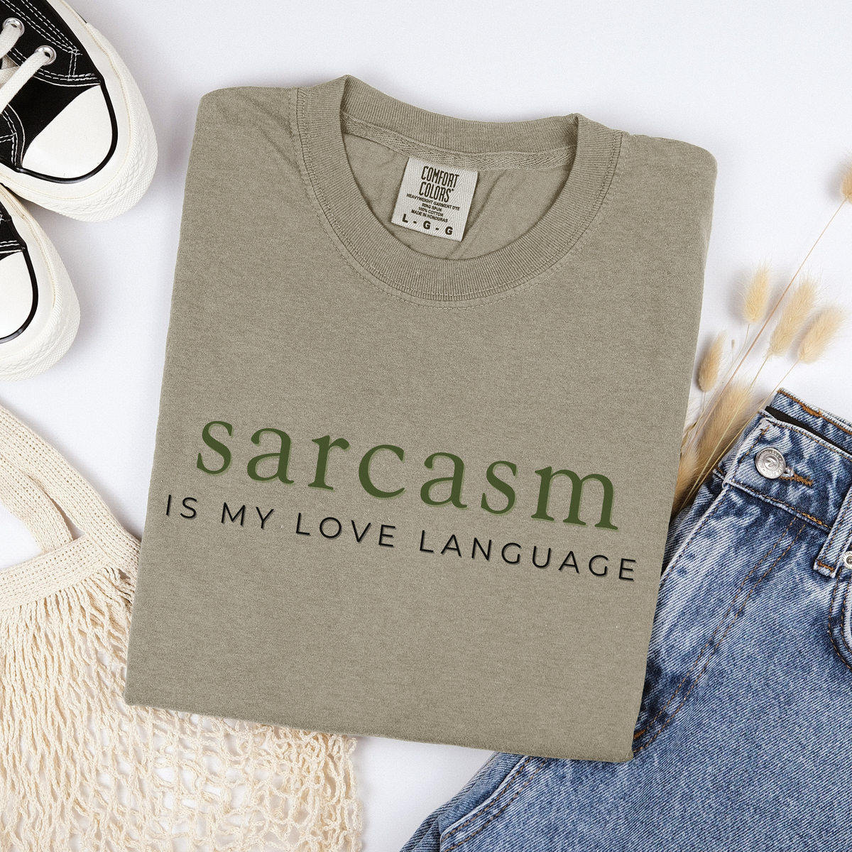 Folded beige t-shirt with 'sarcasm is my love language' text on a white background with jeans and sneakers.