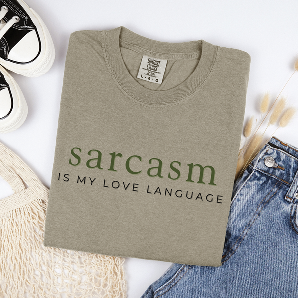 Folded beige t-shirt with 'sarcasm is my love language' text on a white background with jeans and sneakers.