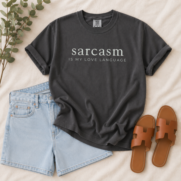 A dark charcoal gray graphic t-shirt with the text 'sarcasm IS MY LOVE LANGUAGE' in white, styled in a flat lay with light blue denim shorts, brown leather slide sandals, and green eucalyptus leaves on a beige fabric background.
