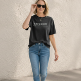Person wearing a black t-shirt with 'sarcasm is my love language' text and blue jeans against a textured wall.