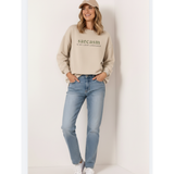 Woman wearing a beige sweatshirt with 'sarcasm is my love language' text and light blue jeans against a white background.