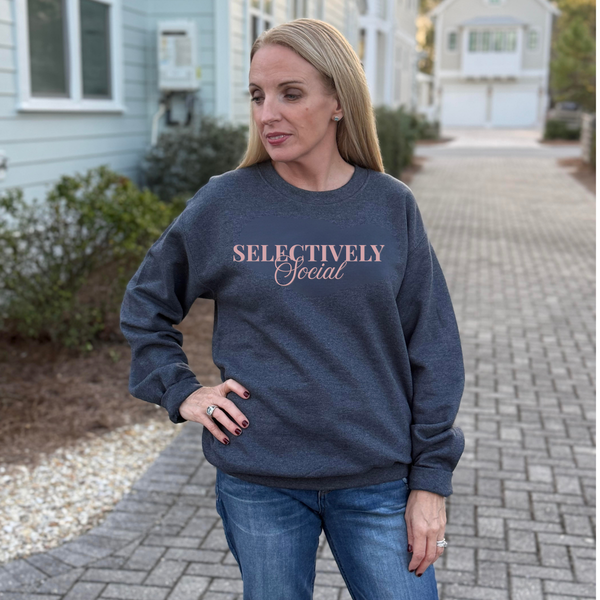 A Woman wearing a dark gray sweatshirt with 'SELECTIVELY Social' text in pink font, standing on a sidewalk.