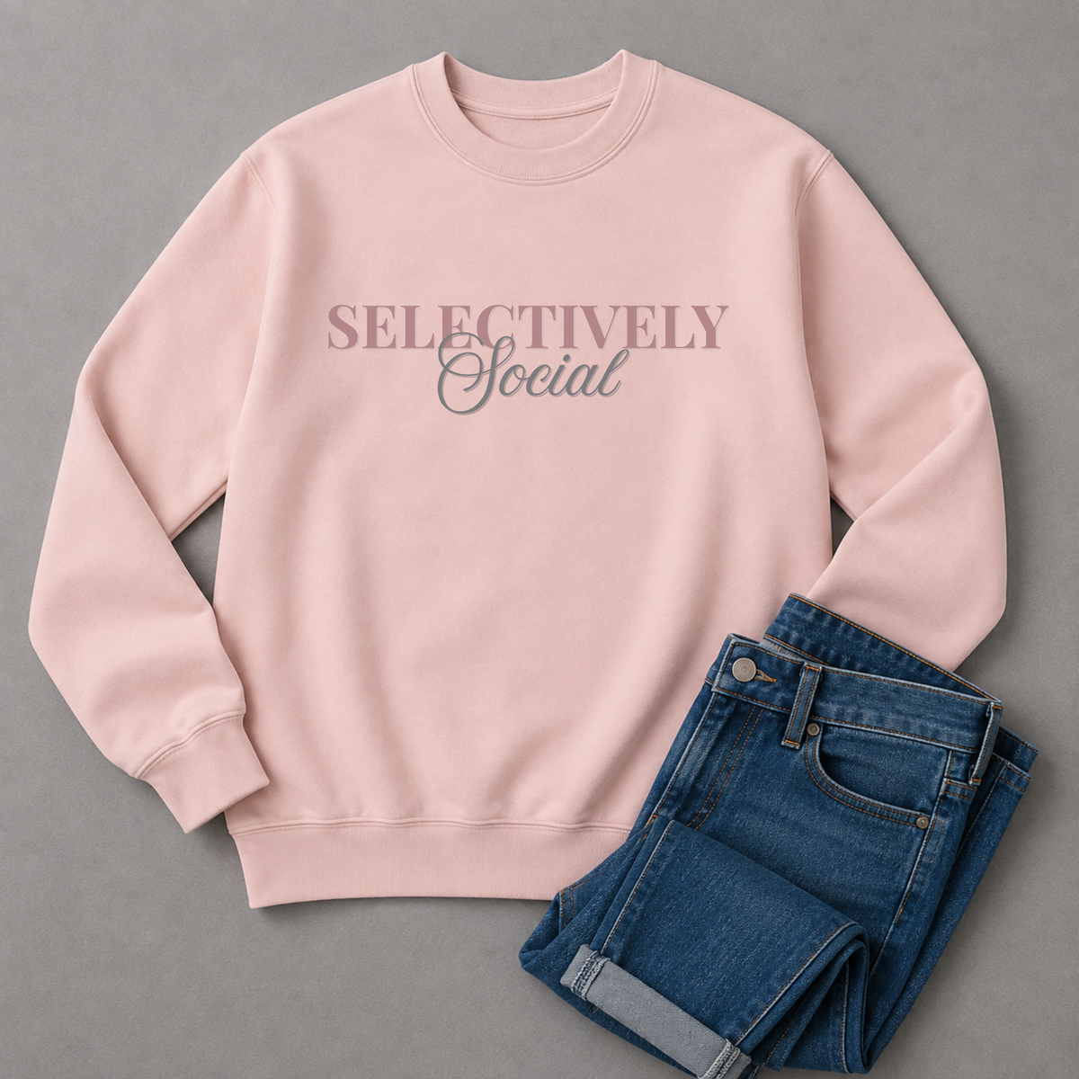 A light pink crewneck sweatshirt with the text 'SELECTIVELY Social' printed across the chest, styled flat-lay with folded blue jeans on a gray background.