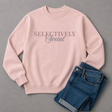 A light pink crewneck sweatshirt with the text 'SELECTIVELY Social' printed across the chest, styled flat-lay with folded blue jeans on a gray background.