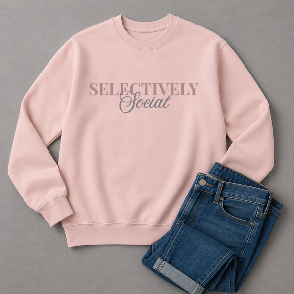 A light pink crewneck sweatshirt with the text 'SELECTIVELY Social' printed across the chest, styled flat-lay with folded blue jeans on a gray background.