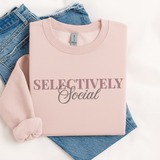 A folded light pink crewneck sweatshirt with the text 'SELECTIVELY Social' printed on the front, styled on top of blue jeans against a white background.