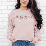 A woman wearing a dusty pink crewneck sweatshirt with the text 'SELECTIVELY Social' printed on the chest, paired with blue jeans, standing against a white brick wall.