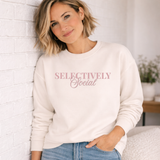 Woman wearing a white sweatshirt with 'SELECTIVELY Social' soft pink text, standing against a light-colored wall.