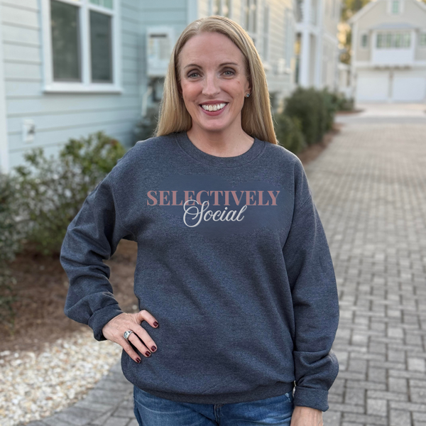 A smiling blonde woman wearing a dark heather gray crewneck sweatshirt with the text 'SELECTIVELY Social' printed on the chest, standing outdoors on a paved walkway with houses in the background.