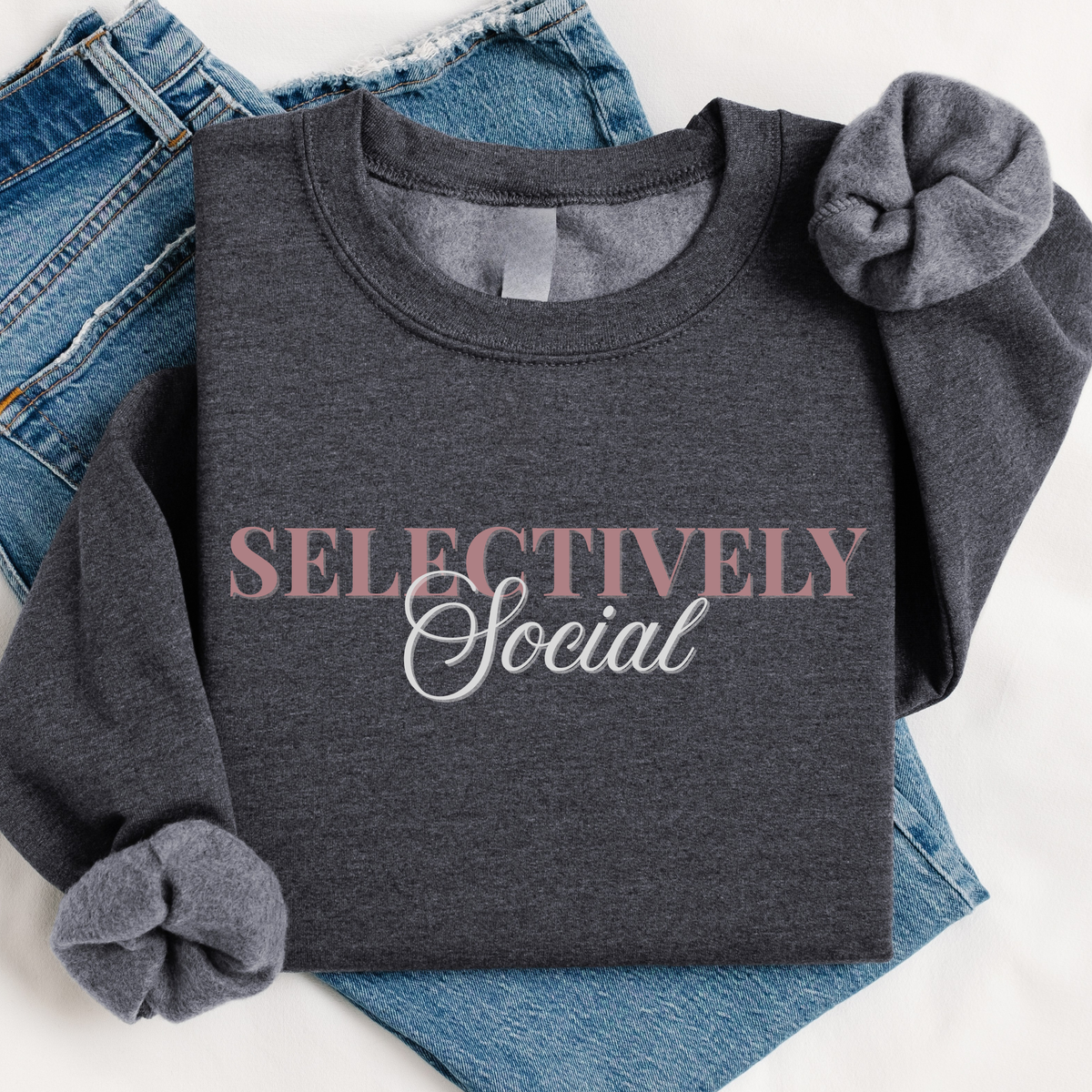 A dark heather gray long-sleeve t-shirt with rolled cuffs, featuring the text 'SELECTIVELY Social' printed on the chest in dusty pink and white. The shirt is styled flat-lay on top of blue denim jeans against a white background.