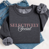 A dark heather gray long-sleeve t-shirt with rolled cuffs, featuring the text 'SELECTIVELY Social' printed on the chest in dusty pink and white. The shirt is styled flat-lay on top of blue denim jeans against a white background.