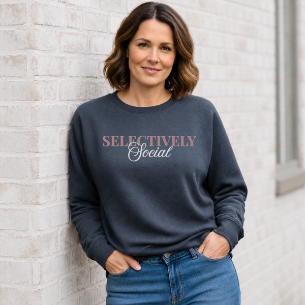 A woman with shoulder-length brown hair stands against a white brick wall wearing a dark charcoal gray sweatshirt with the text 'SELECTIVELY Social' printed on the chest in dusty pink and white, paired with blue jeans.