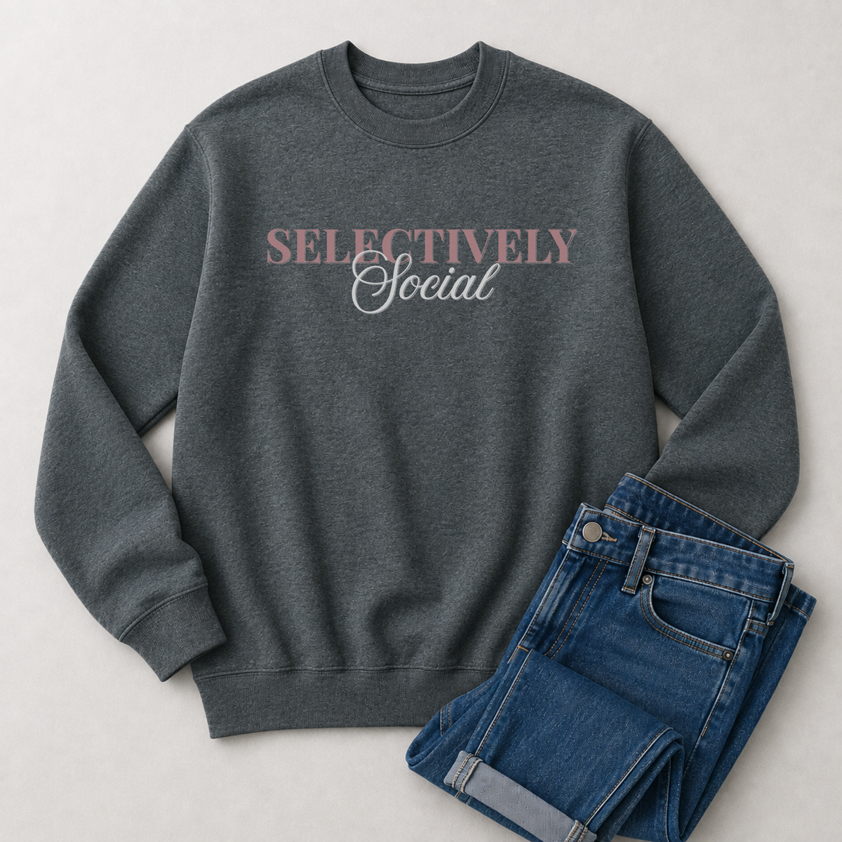 A dark heather gray crewneck sweatshirt with the words 'SELECTIVELY Social' printed across the chest in dusty pink and white, styled flat-lay next to a pair of folded blue jeans on a light gray background.