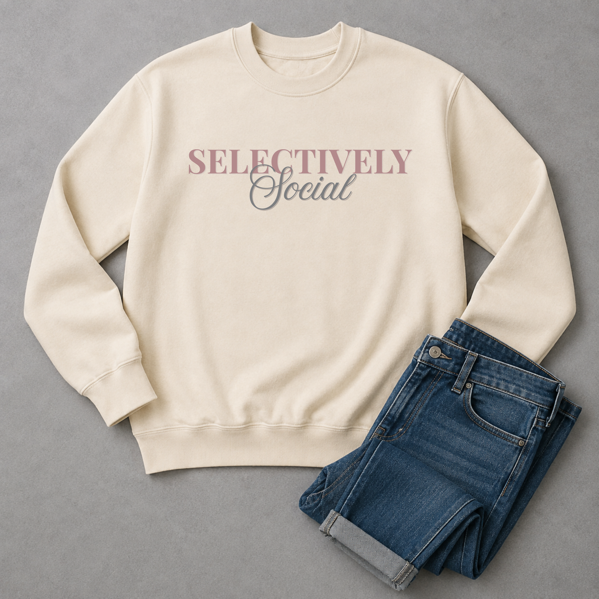 An off-white crewneck sweatshirt with the text 'SELECTIVELY Social' printed on the chest, styled flat-lay next to a pair of folded blue jeans on a gray background.