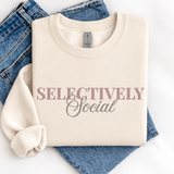 A folded cream sweatshirt with the words 'SELECTIVELY Social' printed across the chest, styled on top of blue jeans against a white background.