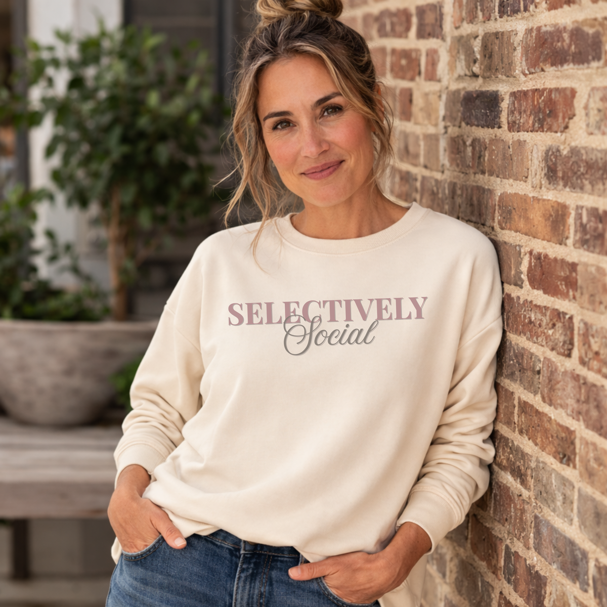 A woman wearing a cream-colored crewneck sweatshirt with the text 'SELECTIVELY Social' printed on the chest, leaning against a brick wall.