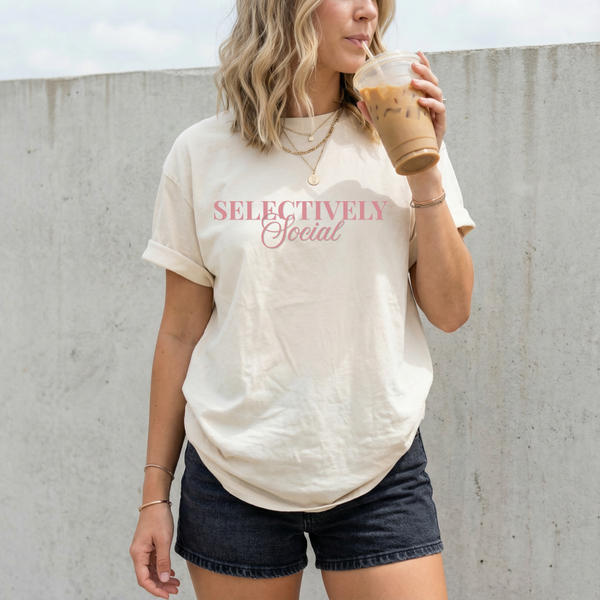 Woman wearing a 'SELECTIVELY Social' t-shirt holding a coffee cup against a concrete wall.