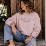 A woman sits on rustic wooden steps wearing a light pink crewneck sweatshirt with the text 'SELECTIVELY Social' in dusty rose and gray, paired with light-wash blue jeans.