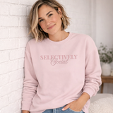 A Woman wearing a pink sweatshirt with 'SELECTIVELY Social' text against a white brick wall.