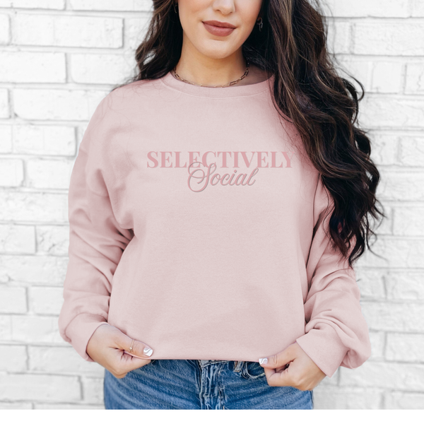 A woman wearing a pink sweatshirt with 'SELECTIVELY Social' text against a white brick wall.