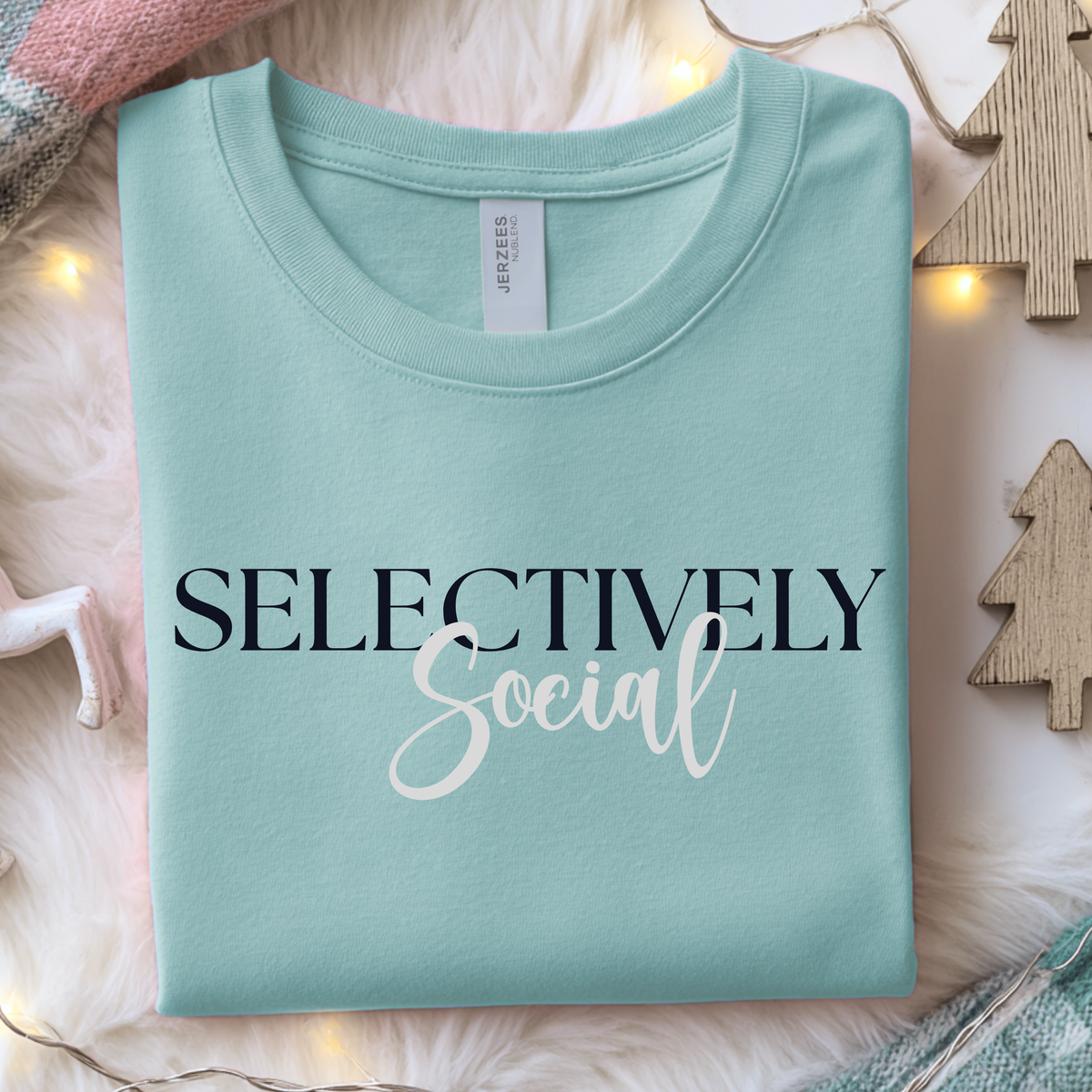 Folded light teal blue sweatshirt with 'SELECTIVELY Social' text on a soft surface with decorative elements.