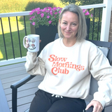 Woman wearing a 'Slow Mornings Club' sweatshirt holding a mug outdoors.