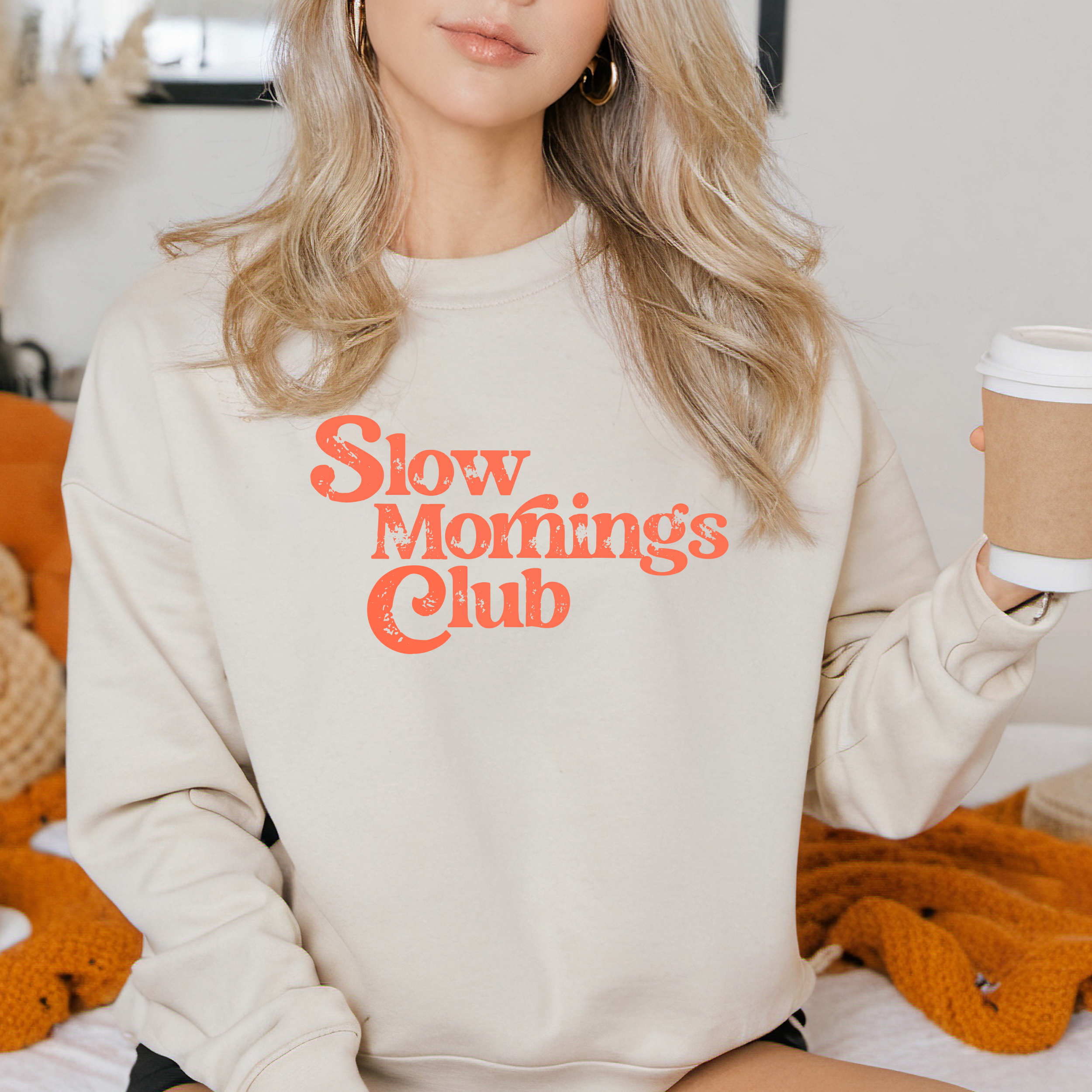 Person wearing a beige sweatshirt with 'Slow mornings club' text, holding a coffee cup.
